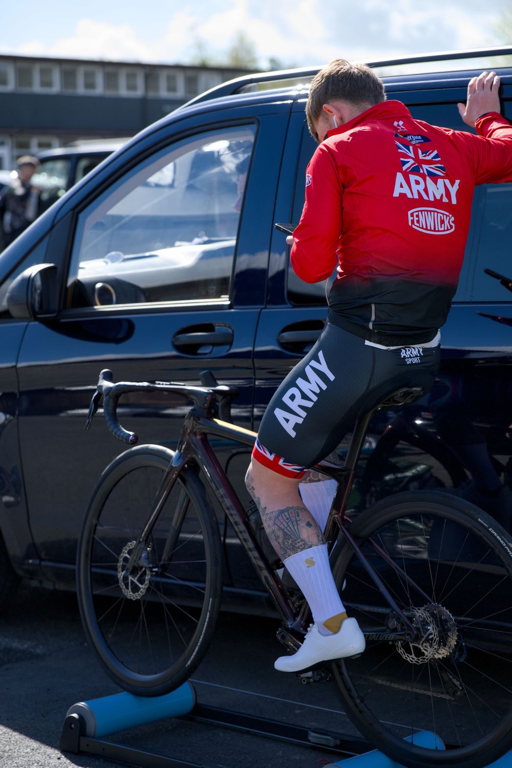 Army cyclist in red jersey warming up on a turbo trainer next to a black van at a cycling event.