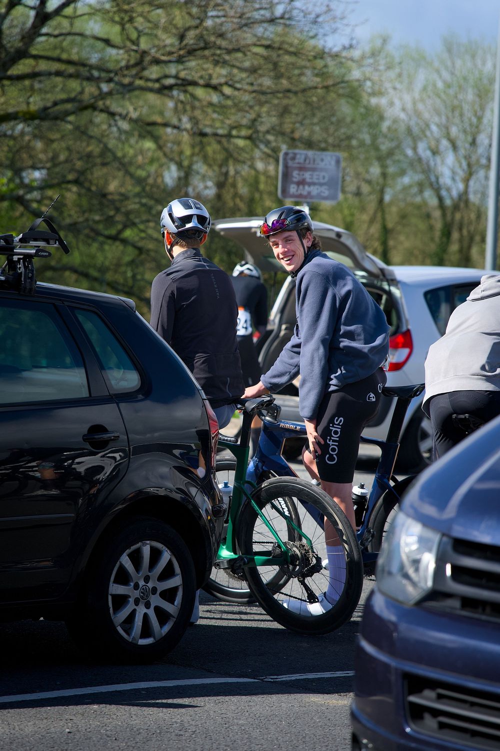 Two cyclists in Cofidis gear stand beside bikes in a parking lot surrounded by cars on a sunny day.