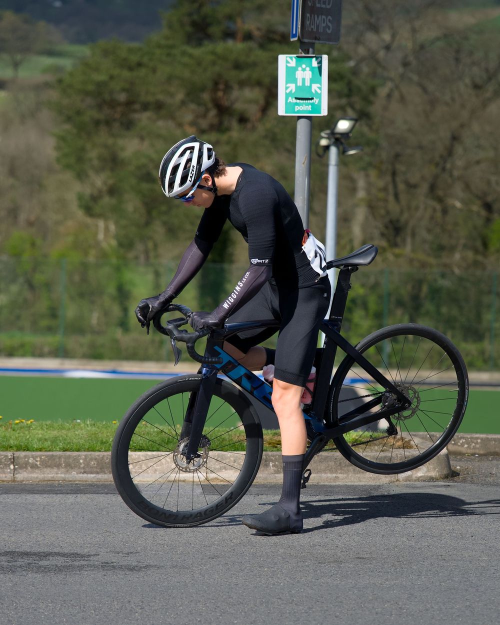 Cyclist in black kit and helmet checks bike on road with green hills in background.