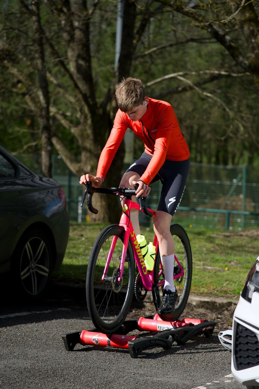 Young cyclist in red jersey trains on pink Specialized road bike using rollers in a sunny outdoor parking area.