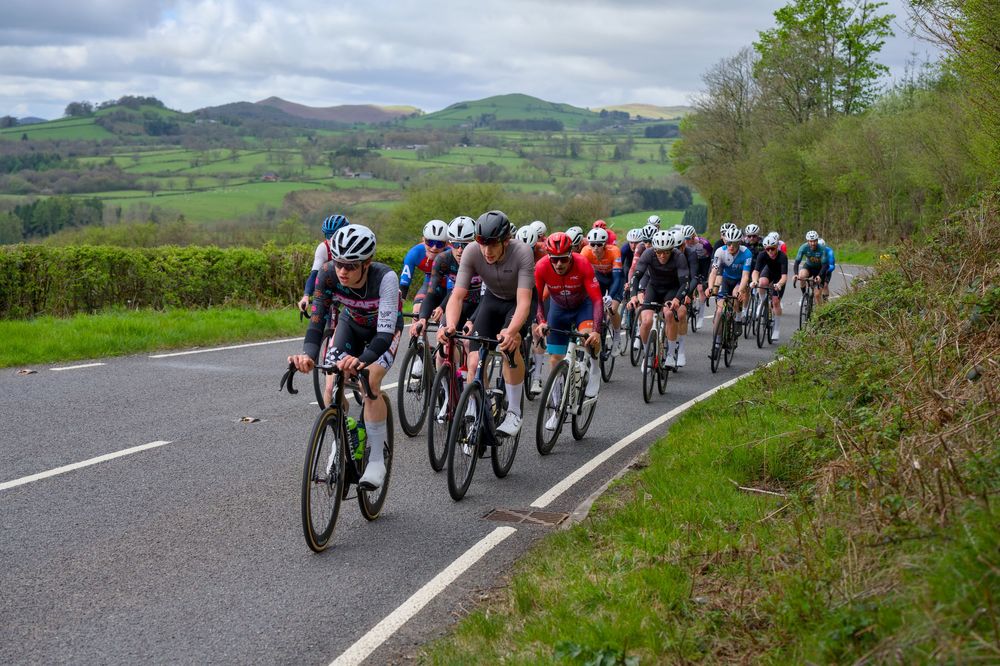 Cyclists racing in a peloton on a rural road surrounded by lush green hills and hedgerows during a cycling event.