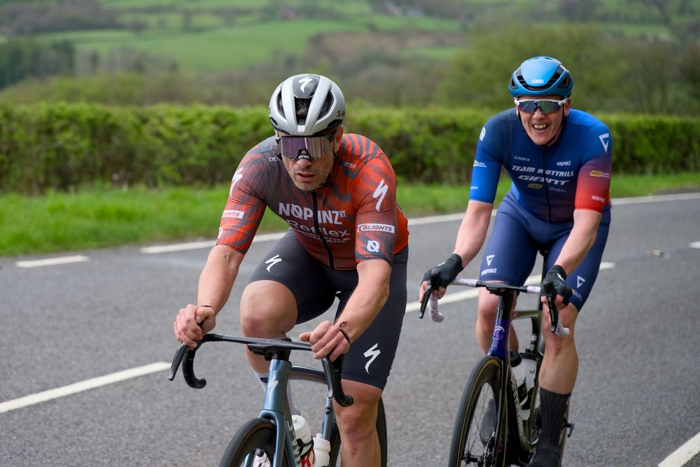 Two cyclists racing on a road, wearing colorful jerseys and helmets, surrounded by green hills.