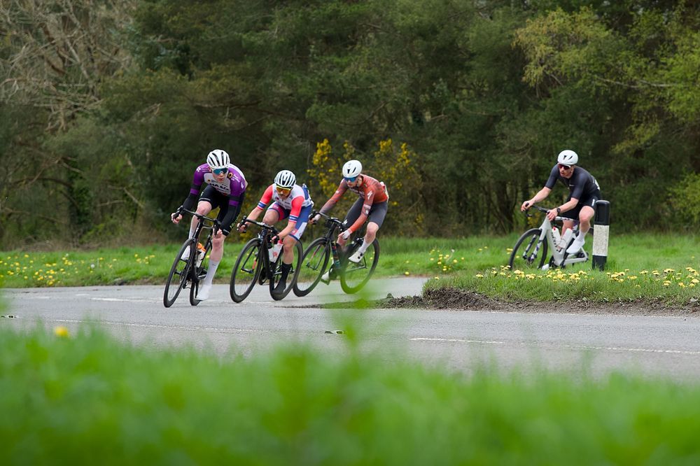 Four cyclists racing on a wet road surrounded by lush green trees and grass during a competitive cycling event.