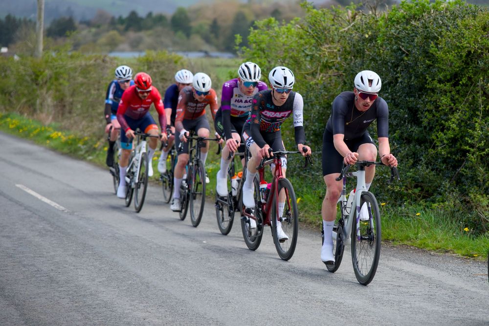 A group of cyclists racing on a narrow country road surrounded by green hedgerows during a road cycling event.