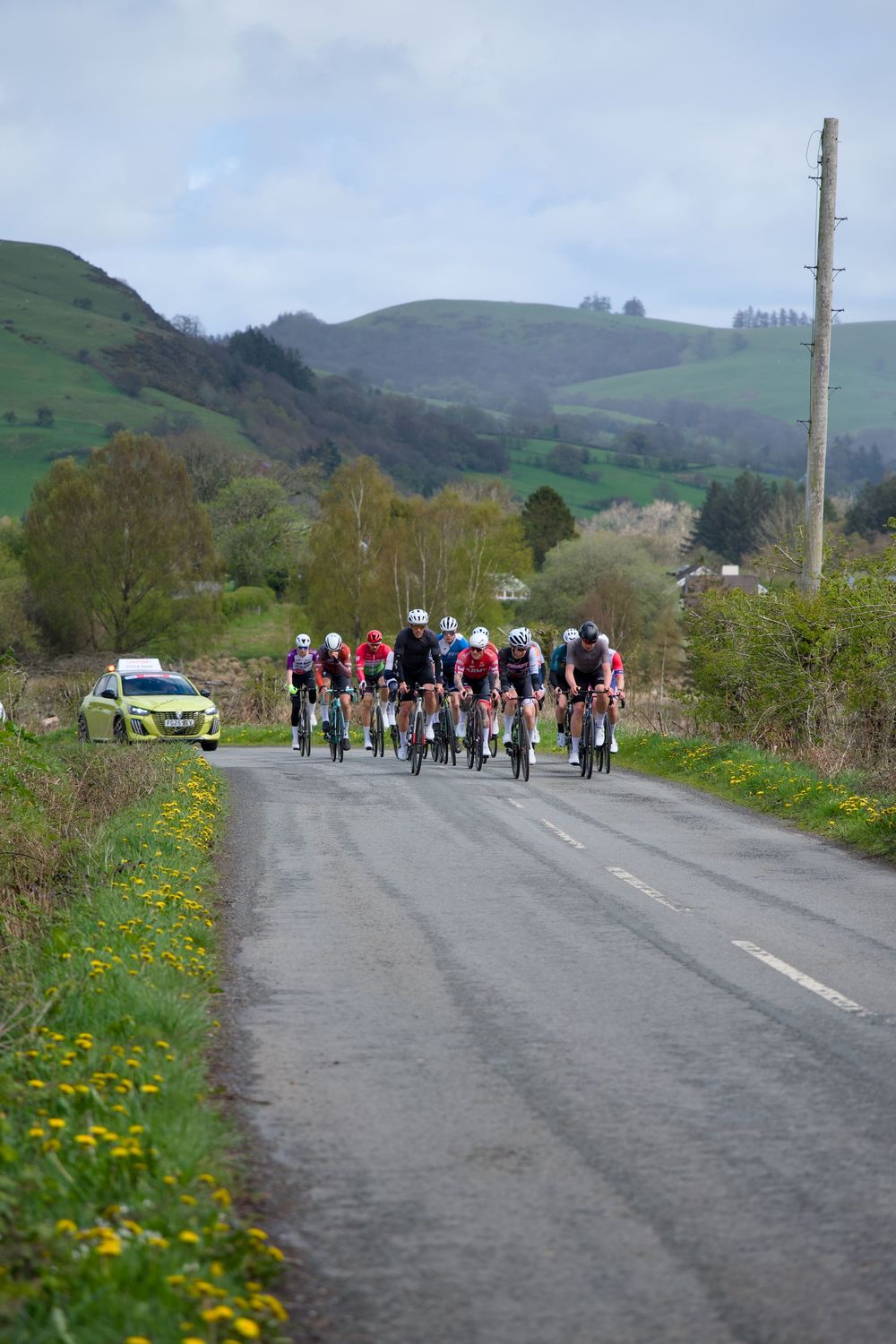 Cyclists race through a rural Welsh countryside road with green hills and yellow wildflowers under a cloudy sky.