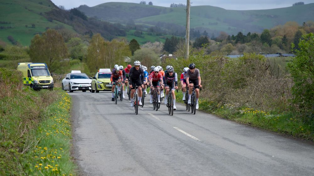 Cyclists competing in a road race on a rural road with support vehicles and scenic hilly countryside in background.