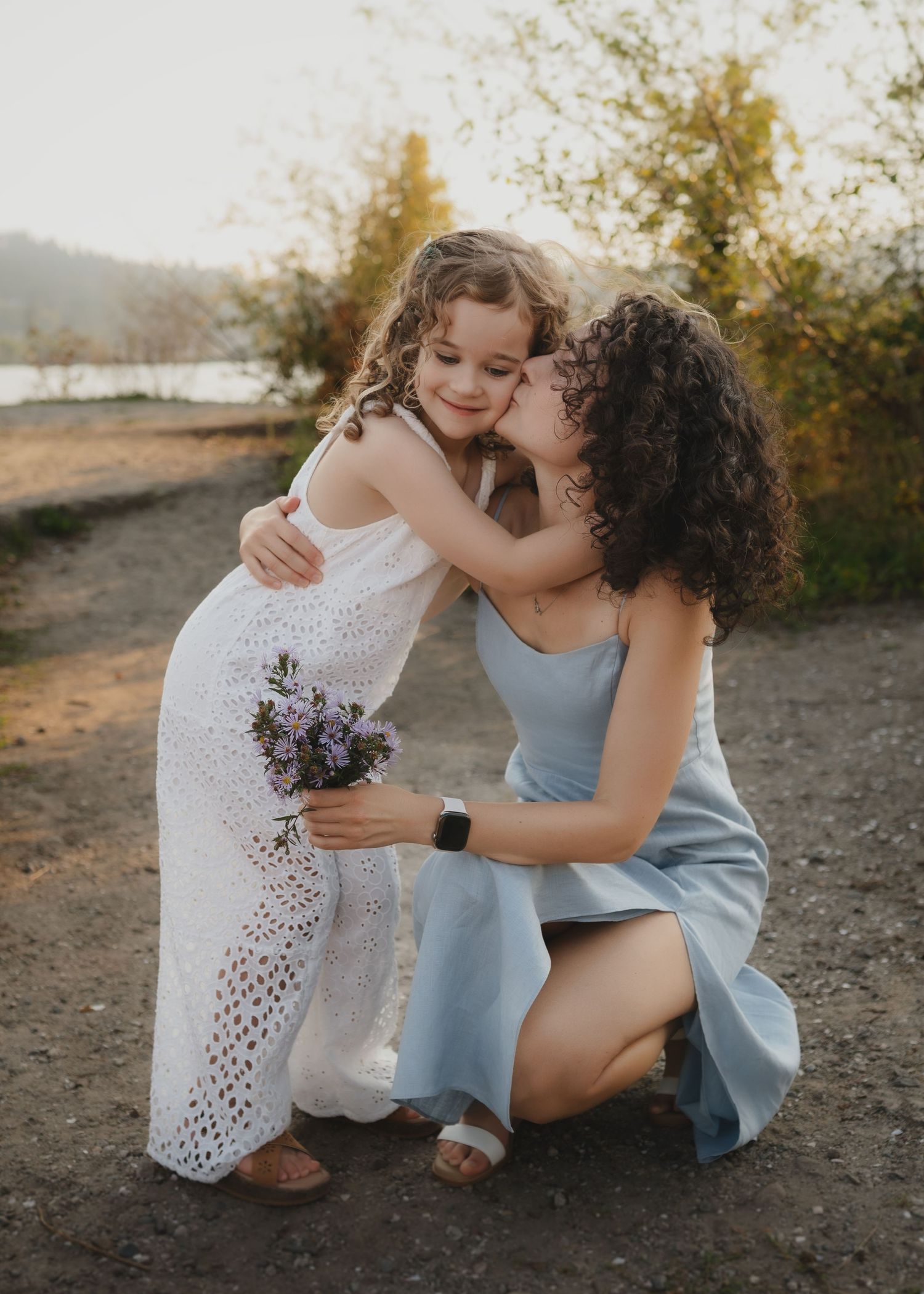 A woman kneeling outdoors kisses a young girl in a white dress holding purple flowers near a lake.
