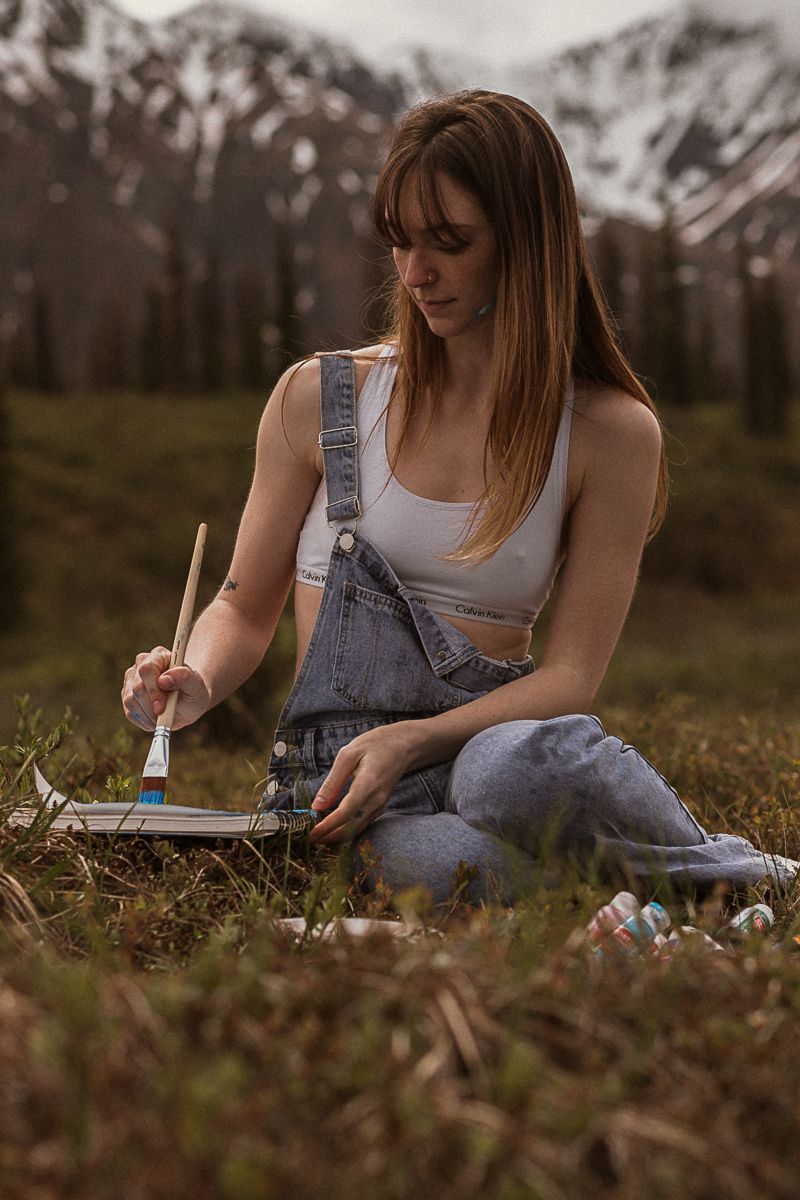 skinny woman in overalls and white bra painting in a field with snowy mountains behind her