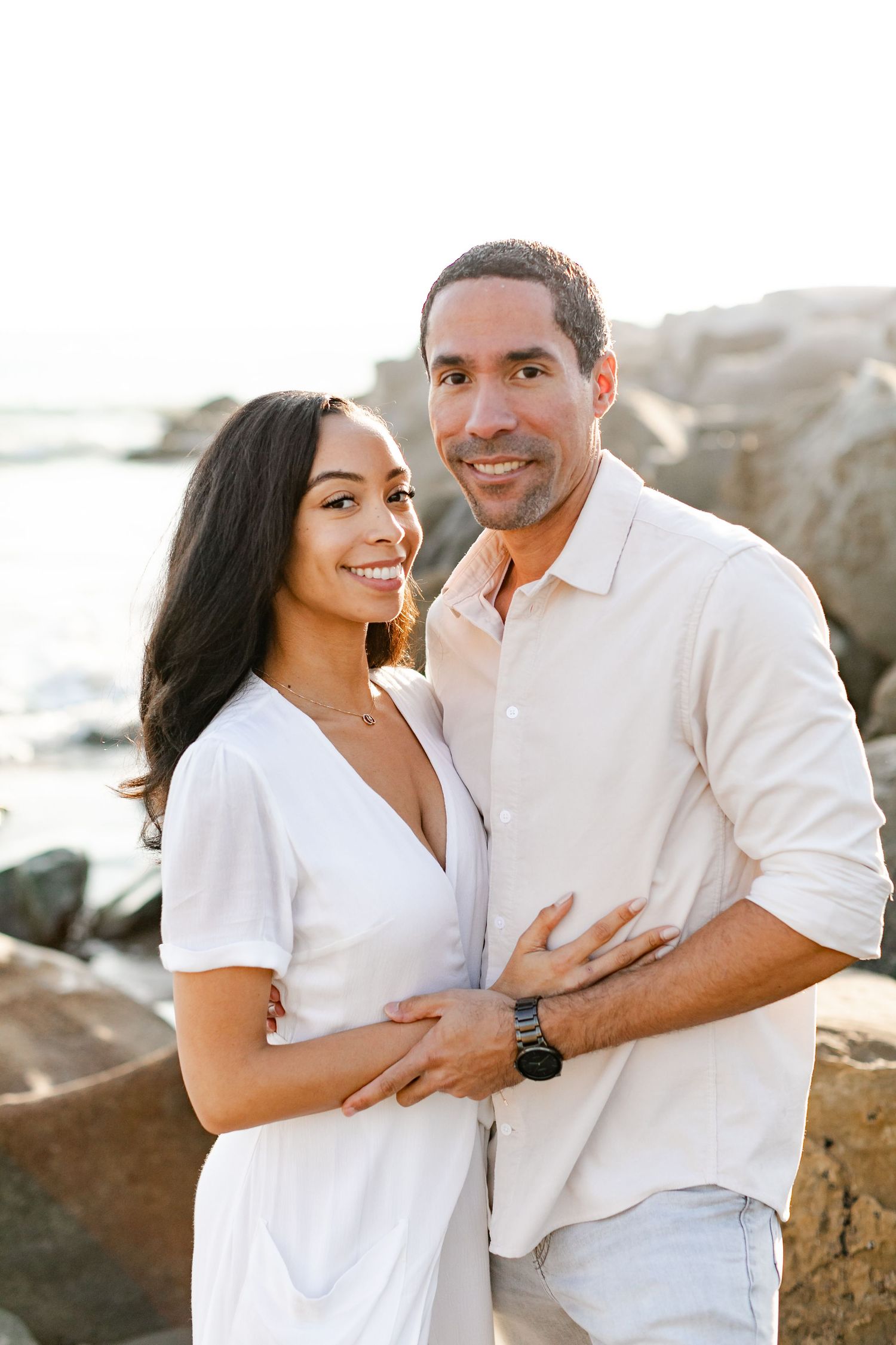 Smiling couple embracing on rocky beach at sunset during outdoor engagement photo session.