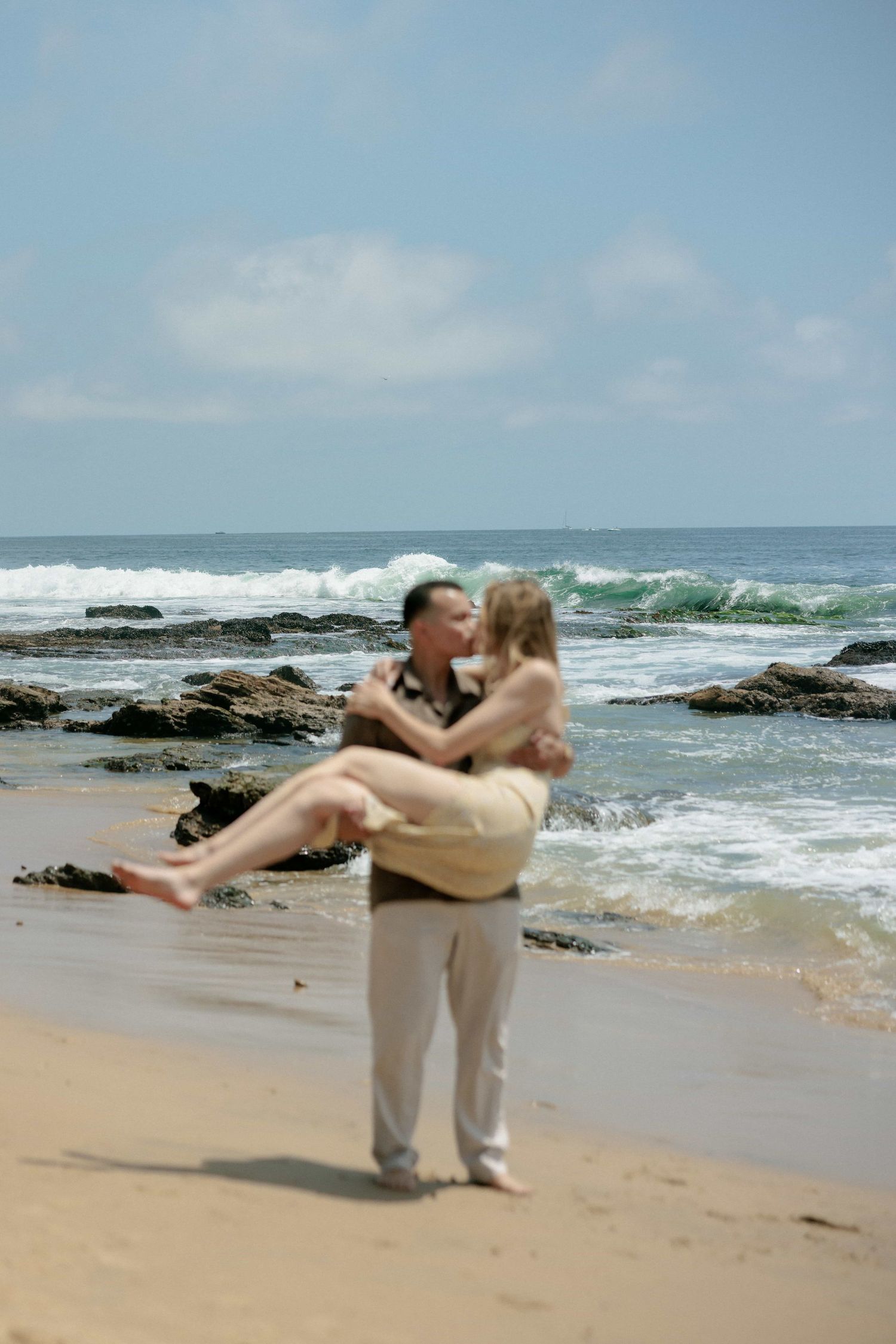Man carrying woman in his arms while kissing on a rocky ocean beach.