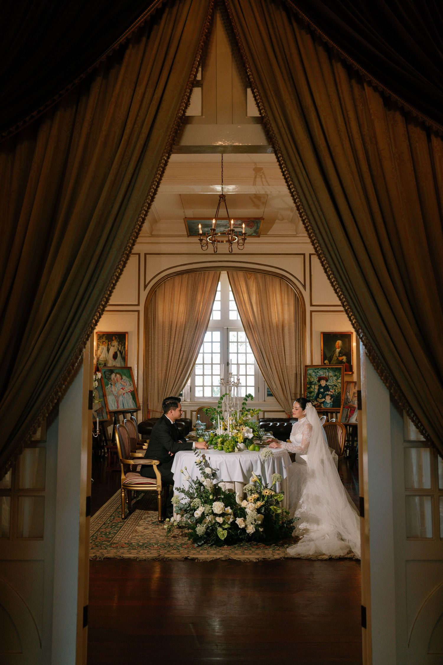 Bride and groom seated at elegant reception table framed by dramatic draped doorway with chandelier.
