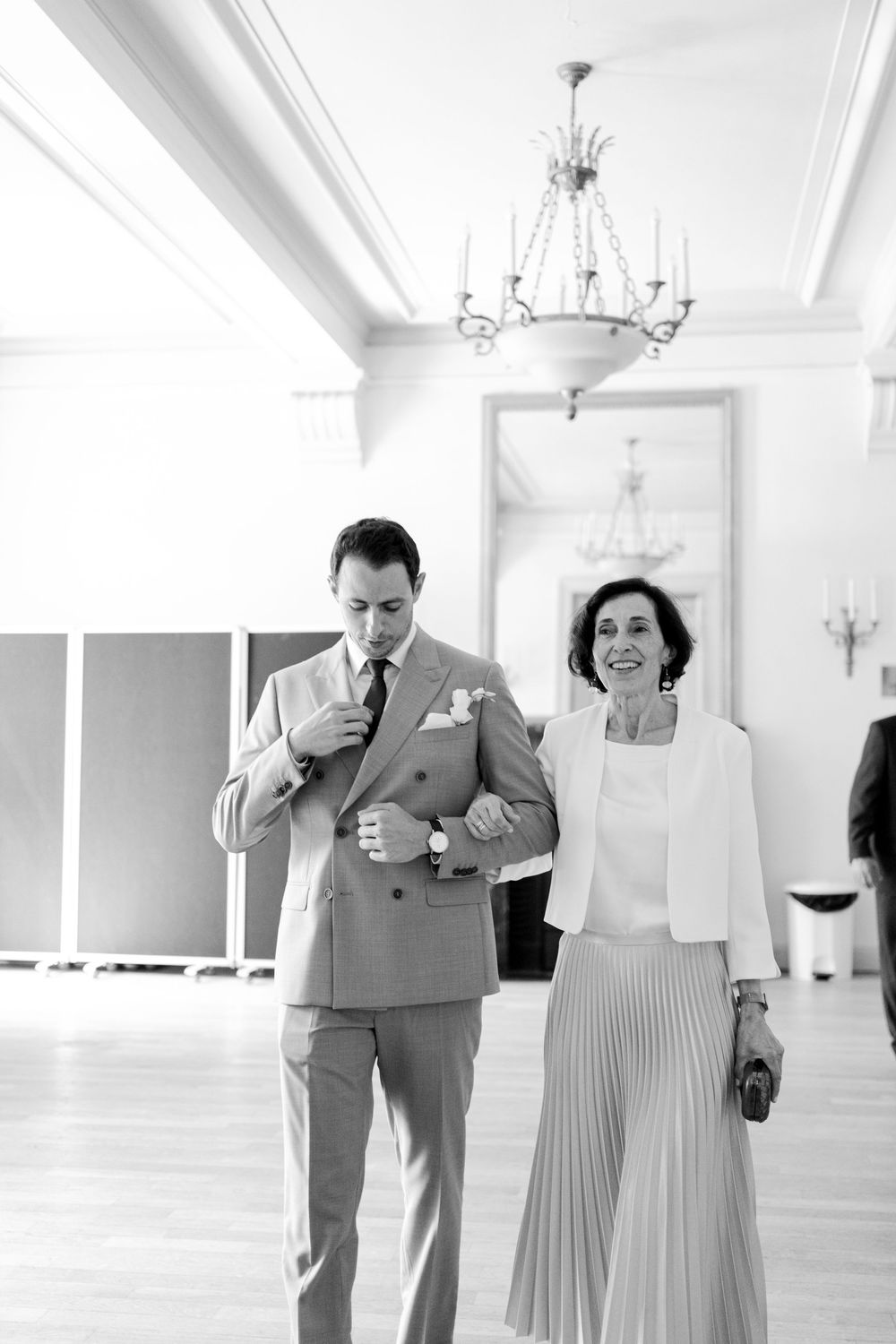 Groom in gray suit adjusting jacket while walking with elegant woman in white blazer in a grand ballroom.