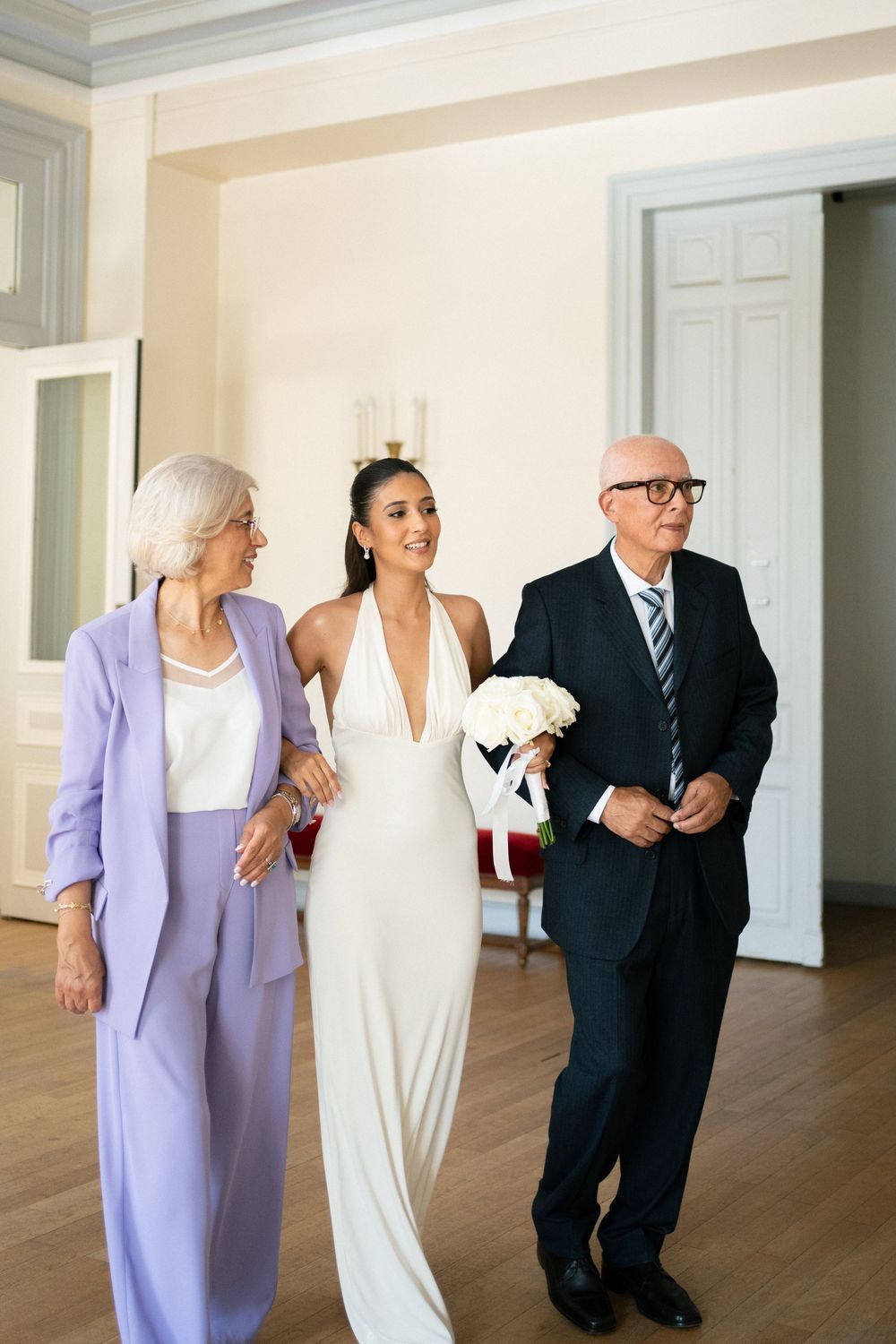 Bride in white halter gown walks with elderly parents in elegant venue, holding white rose bouquet.