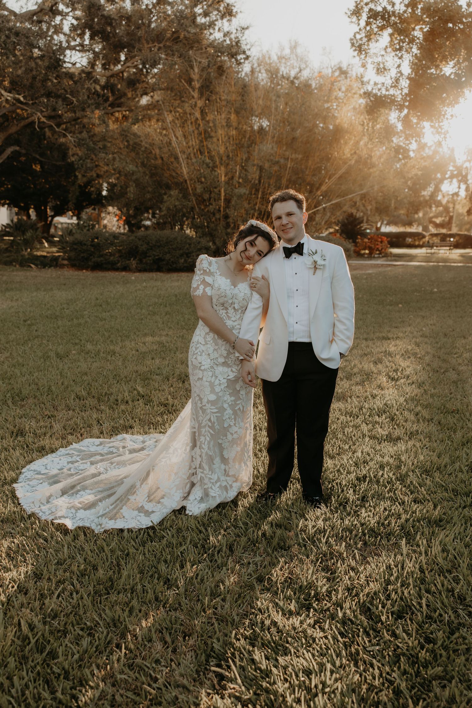 Bride and groom posing together on green lawn at golden hour, she in lace gown with train, he in white tuxedo jacket.
