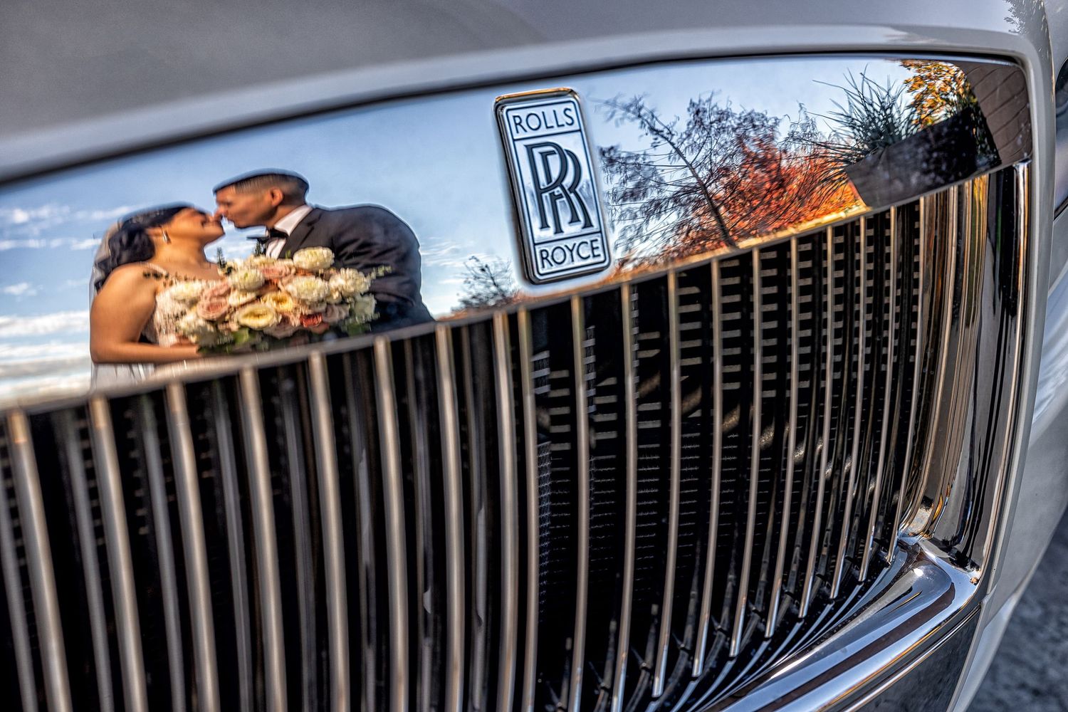 A creative wedding portrait of a bride and groom kissing, perfectly reflected in the chrome grille of a vintage Rolls-Royce luxury car.