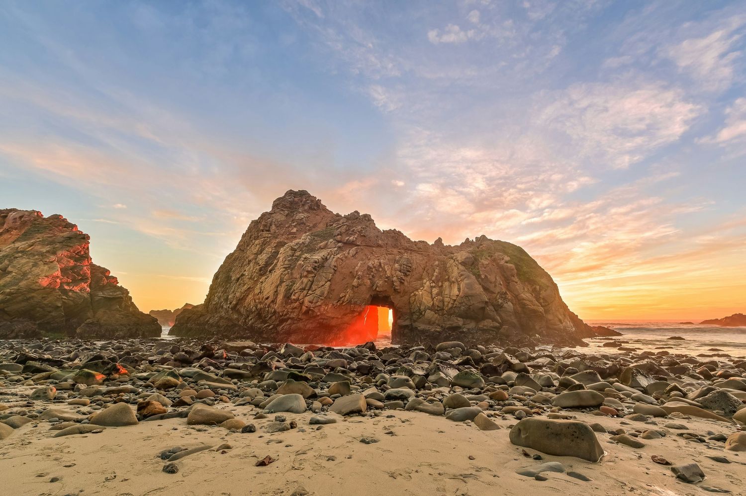 Stunning sunset through a natural rock arch on a rocky beach with glowing orange light and colorful sky.