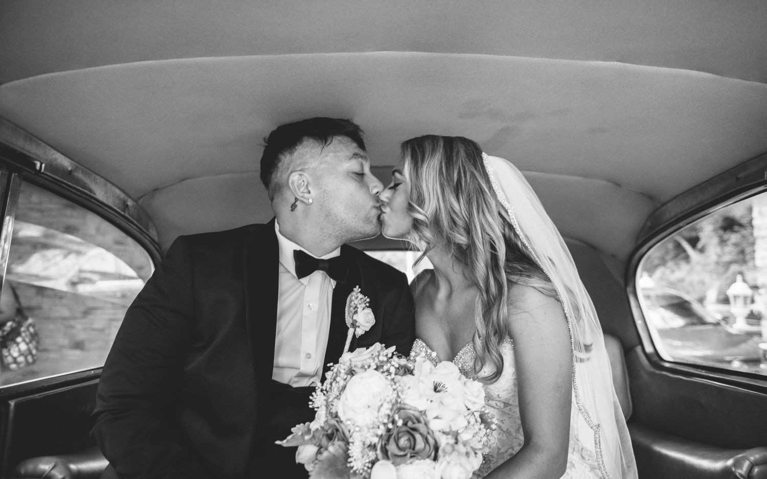 A bride and groom share a tender kiss in a vintage car in a black and white wedding photo.