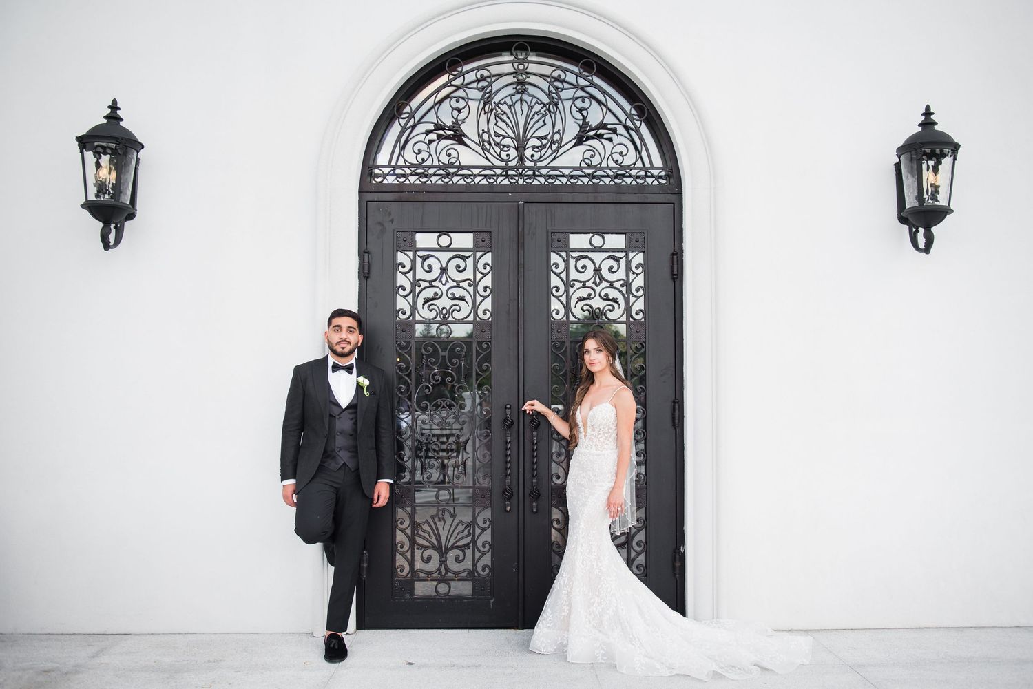 Bride and groom pose before ornate black iron doors with decorative lanterns on a white building.