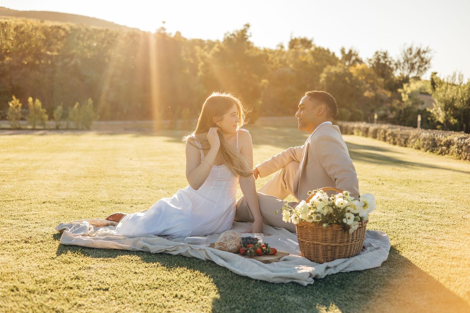 Couple enjoying a romantic sunset picnic on green grass with a wicker basket of flowers in warm golden light.