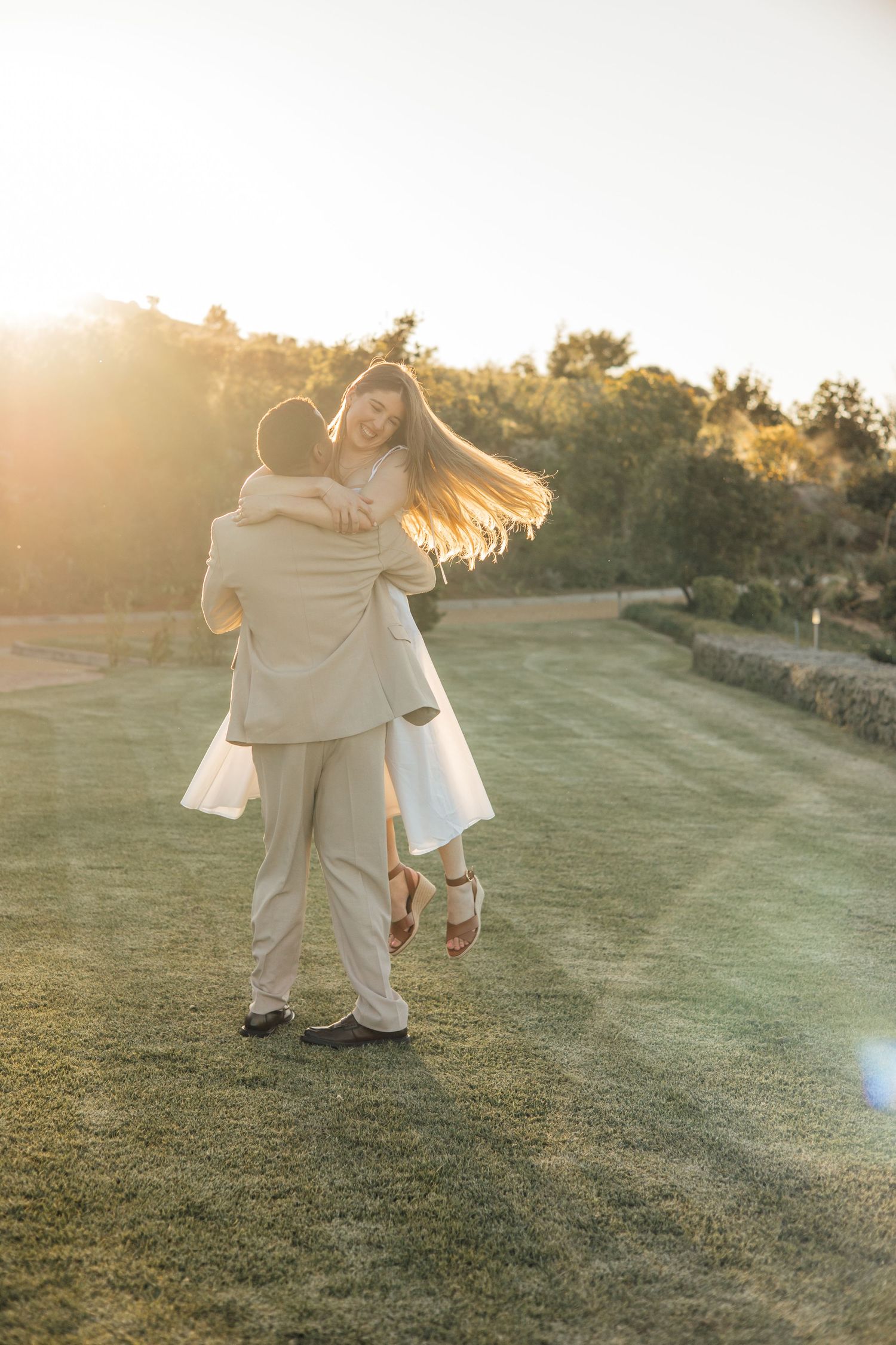 A joyful couple embraces at sunset on a lush green lawn, the groom lifting his bride in a golden backlit glow.