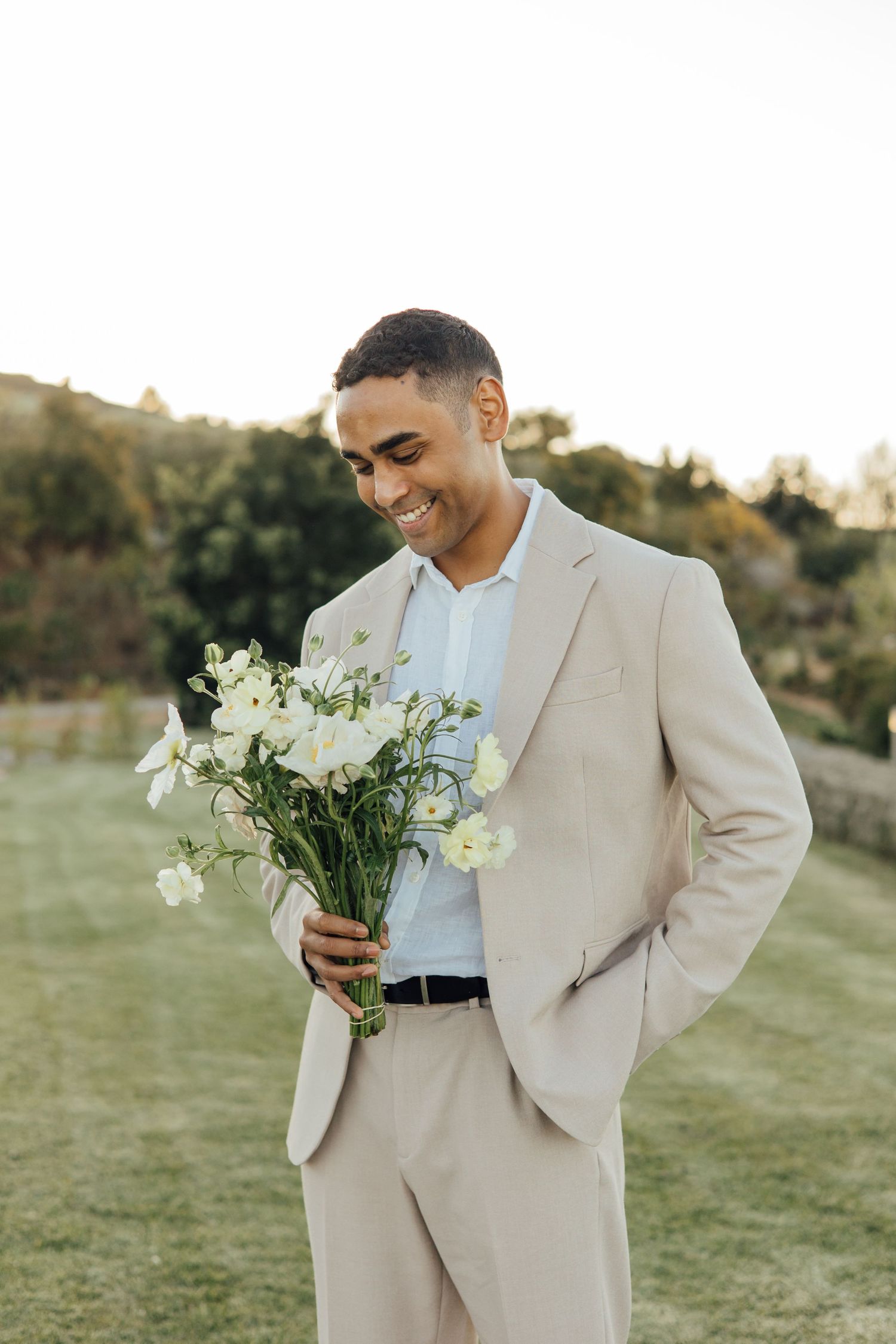 Smiling man in beige suit holding white flower bouquet outdoors at golden hour.