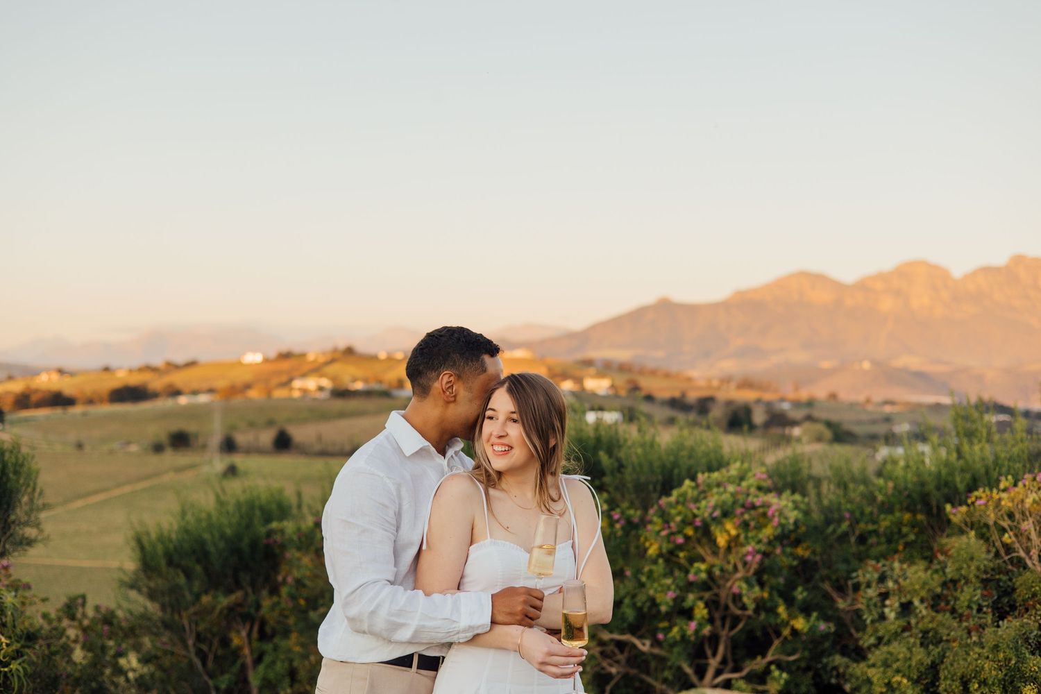 Couple sharing a romantic moment at sunset with scenic vineyard and mountain views, holding champagne glasses.