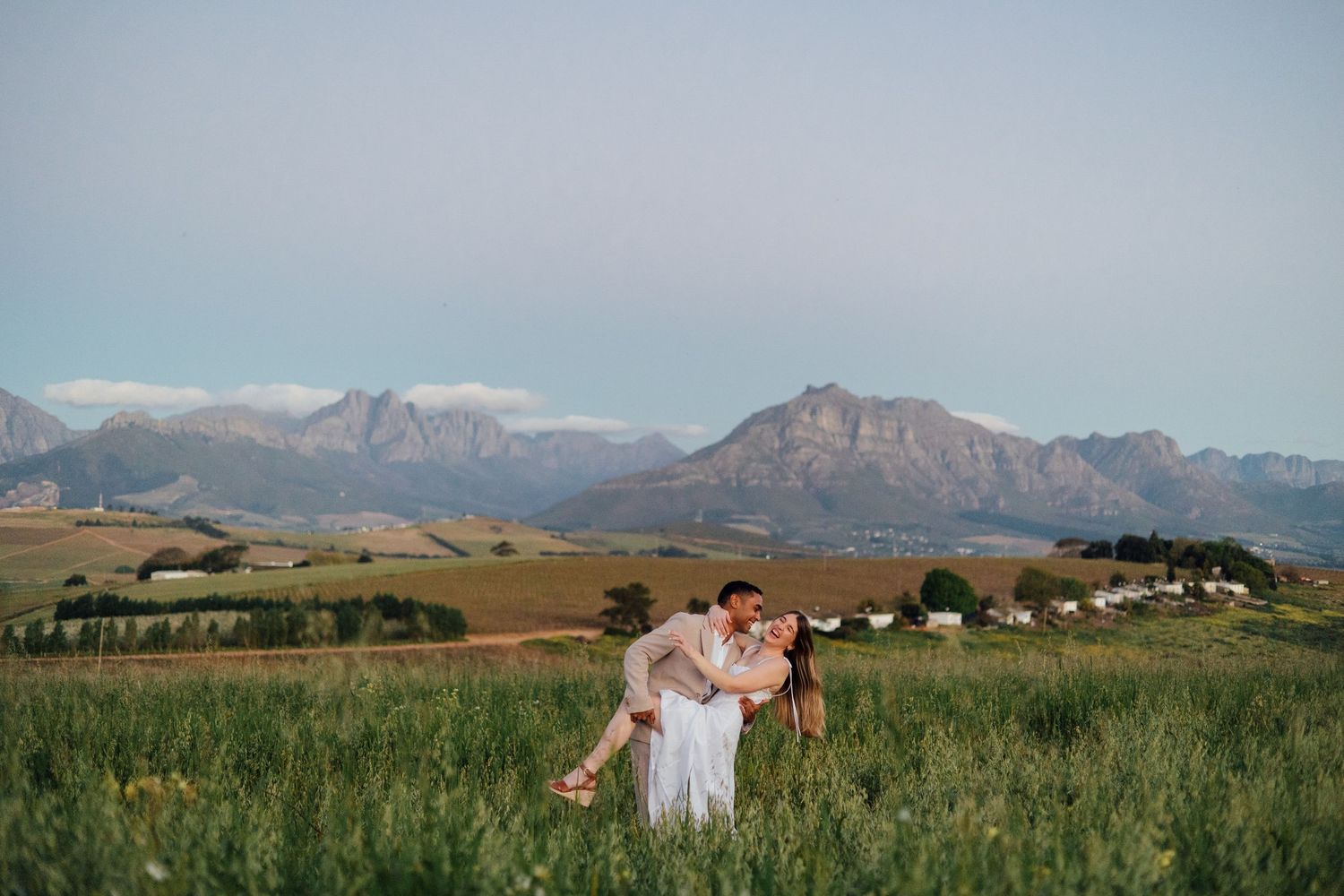 Couple embracing in lush green field with stunning mountain backdrop during golden hour in South Africa.