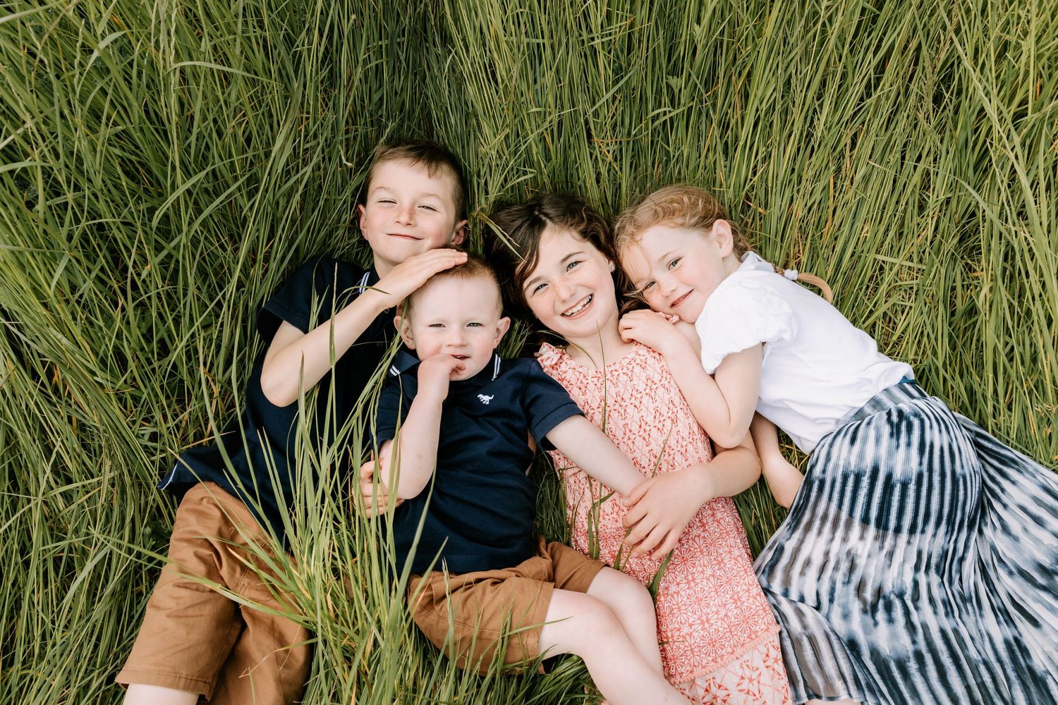 Four children lying in tall green grass, smiling and hugging each other in a joyful outdoor family portrait. Family Photographer Waterford