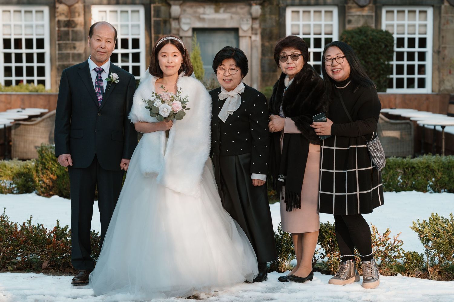 A bride in a white gown poses with family in a snowy winter garden outside a stone building.