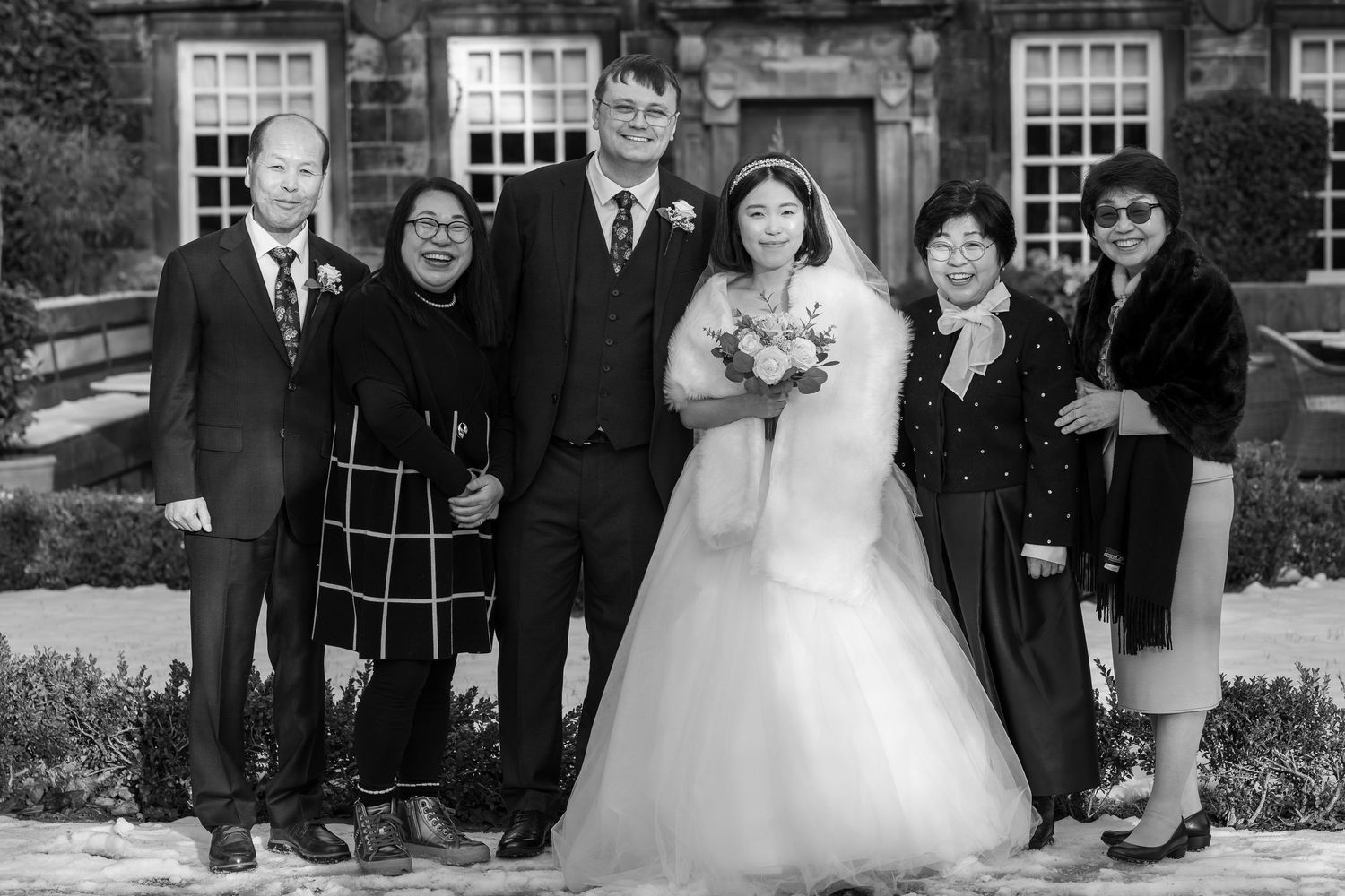 Black and white wedding photo of bride and groom with family posing outdoors in snow near a stone building.