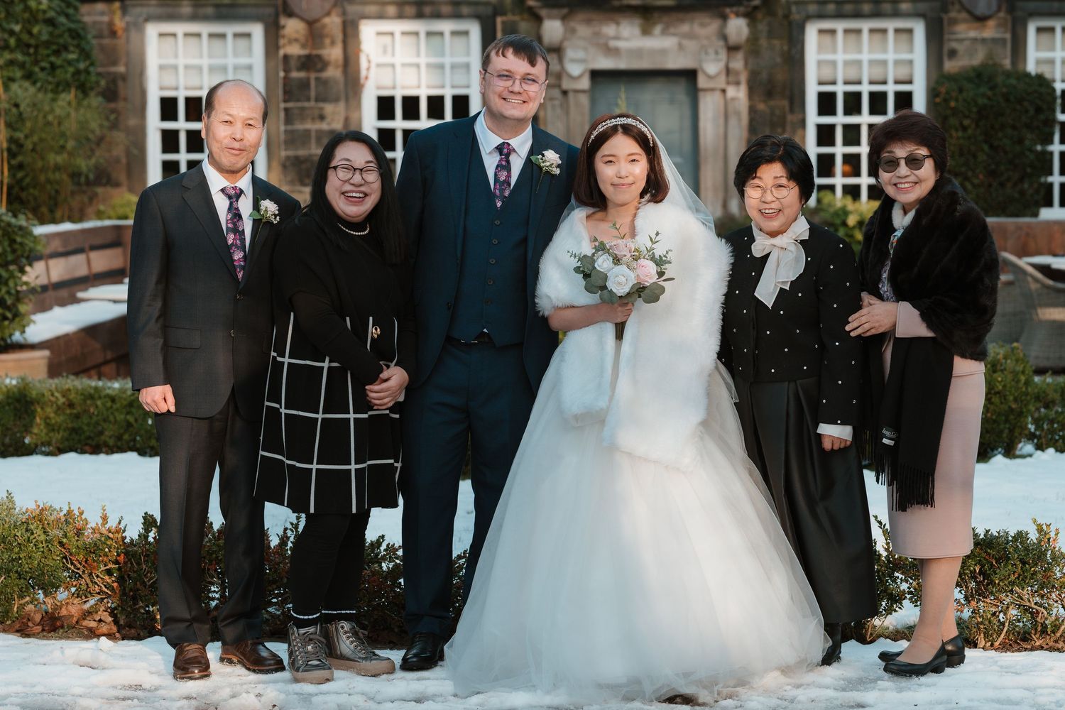 Wedding couple poses with family in snow outside stone building, bride in white gown holding bouquet, winter ceremony.