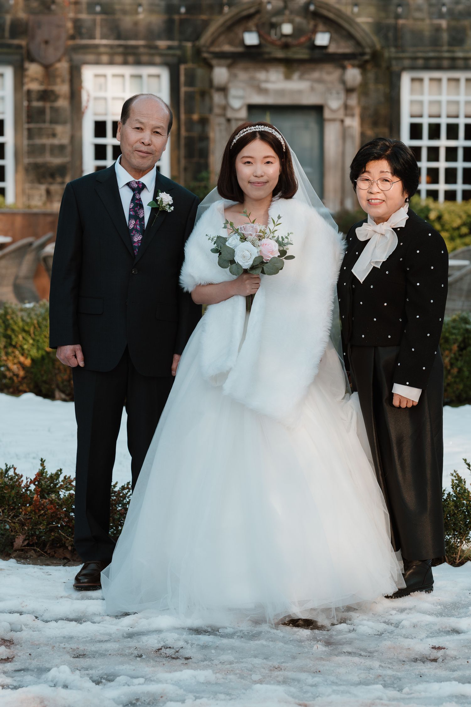 Bride in white gown with bouquet poses with two family members in snow outside historic stone building.