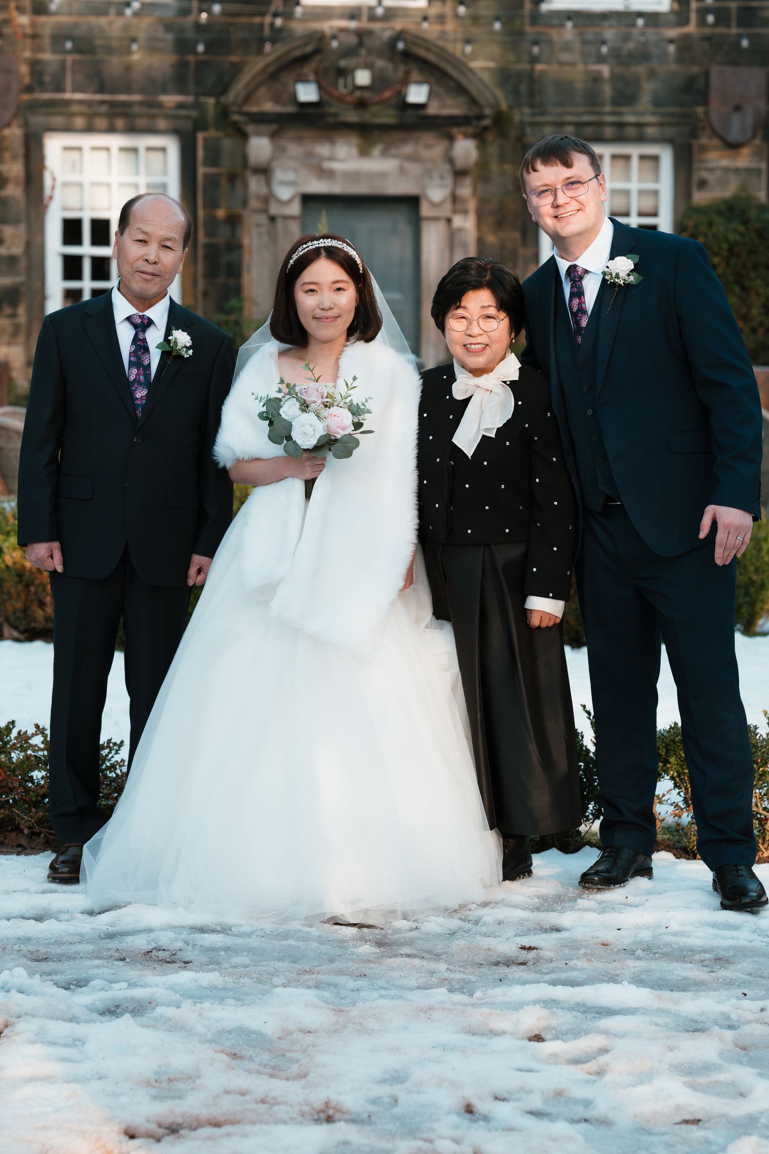 A bride in a white gown poses with three family members outside a snow-covered estate during a winter wedding.