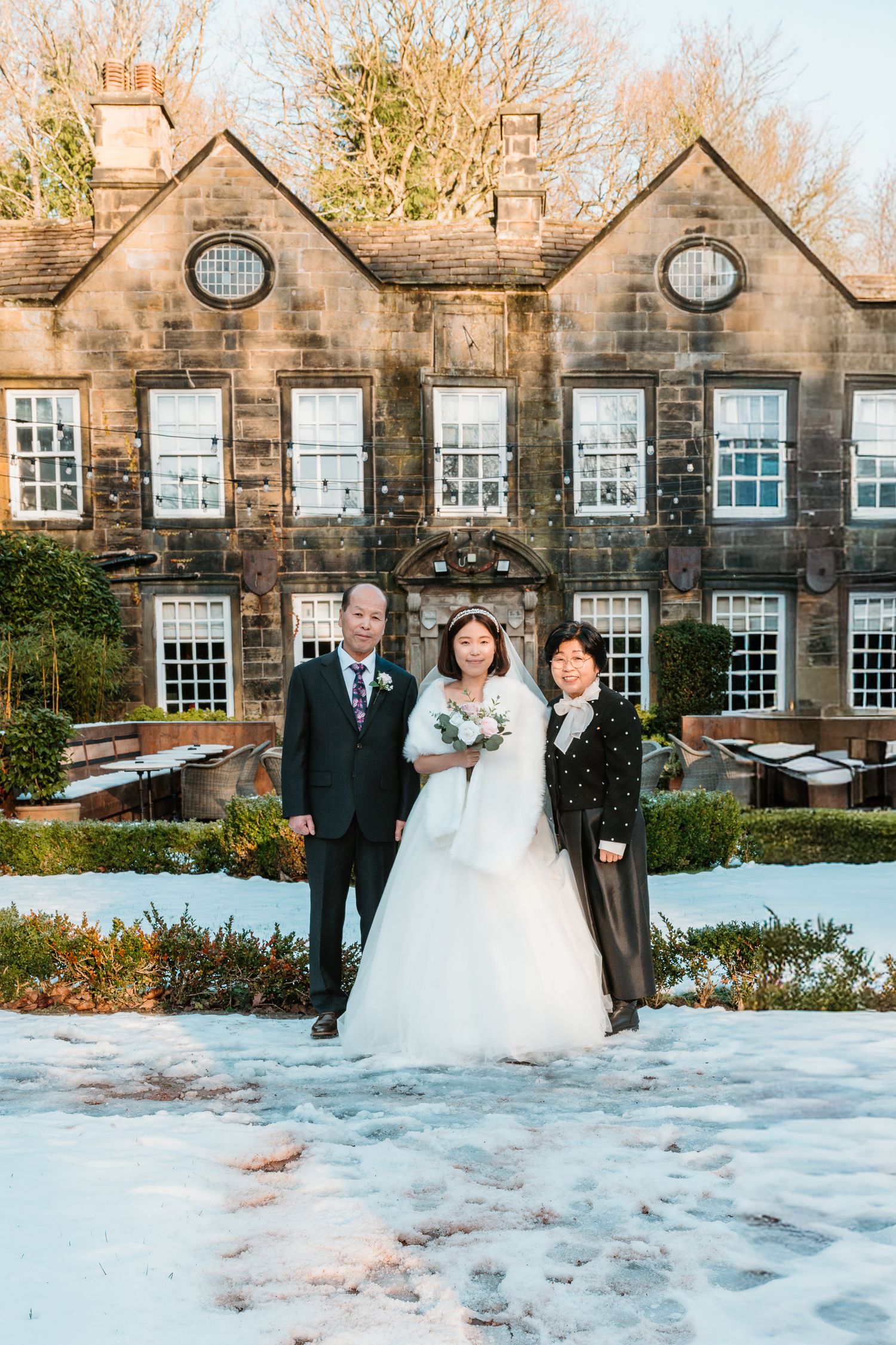 Bride in white gown with family posing in snow outside a stone manor house venue on a winter wedding day.