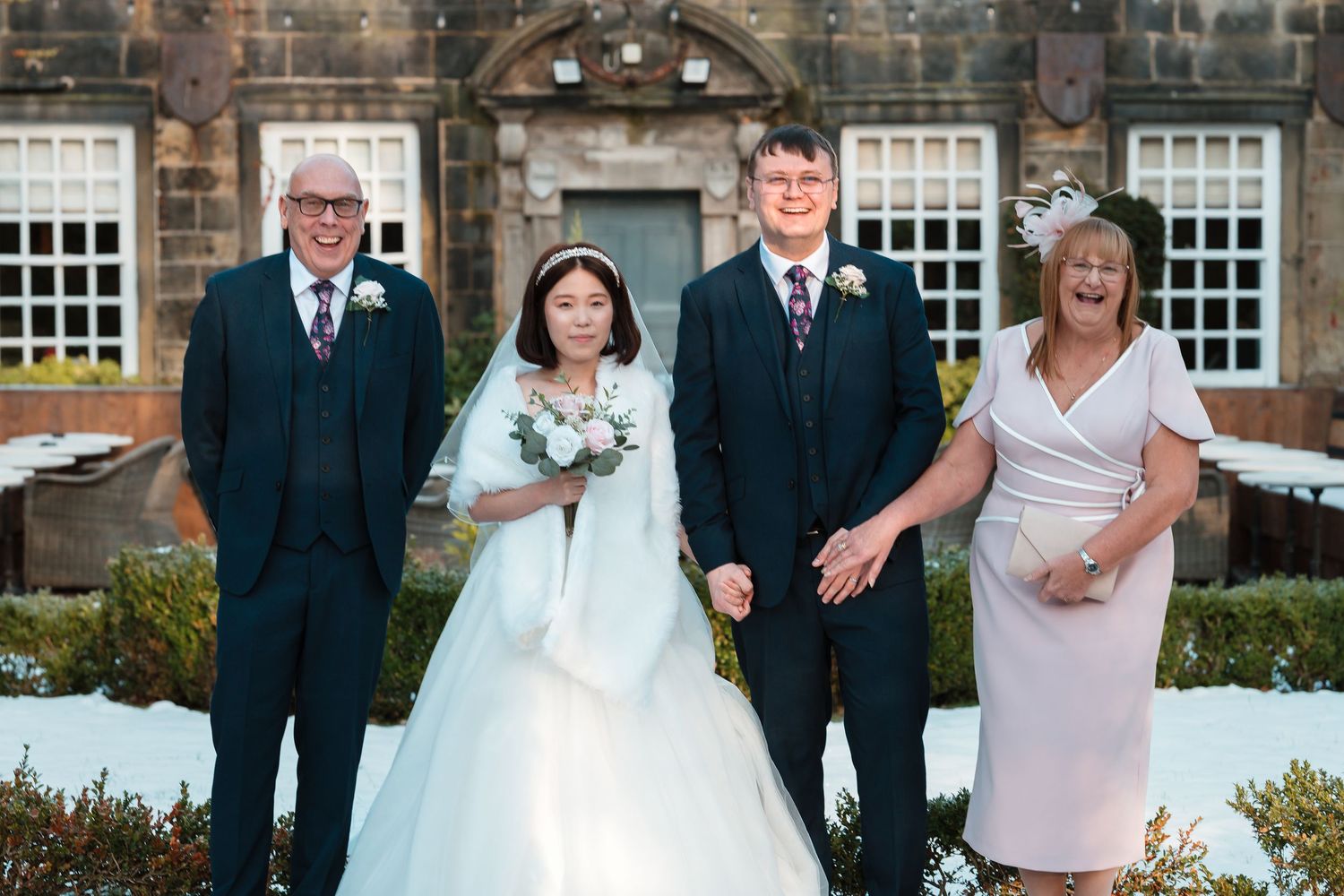 Wedding party of four posing outdoors in snow, bride in white gown with bouquet flanked by family in formal attire.