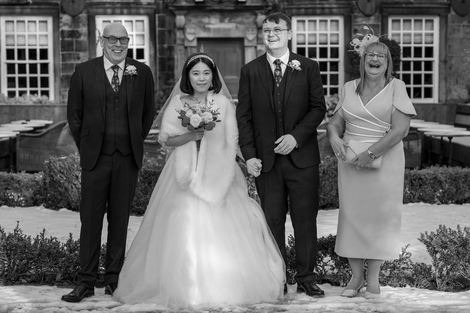 Black and white wedding photo of bride in gown with bouquet, groom, and two guests posing outside historic venue.