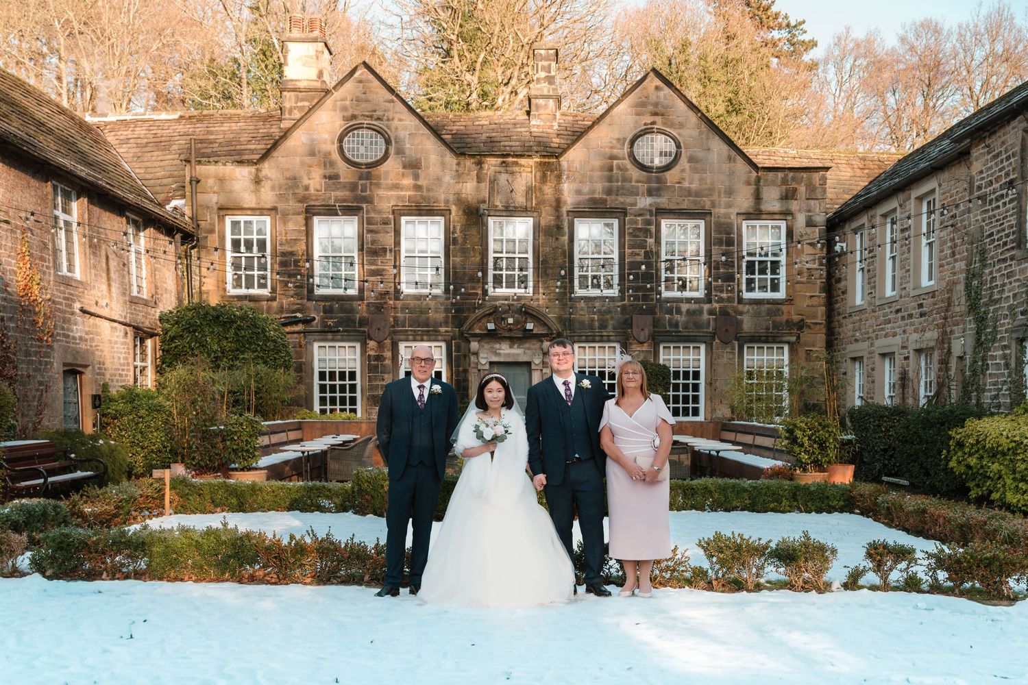Wedding party of four posing in snow outside a grand stone manor house in winter sunshine.