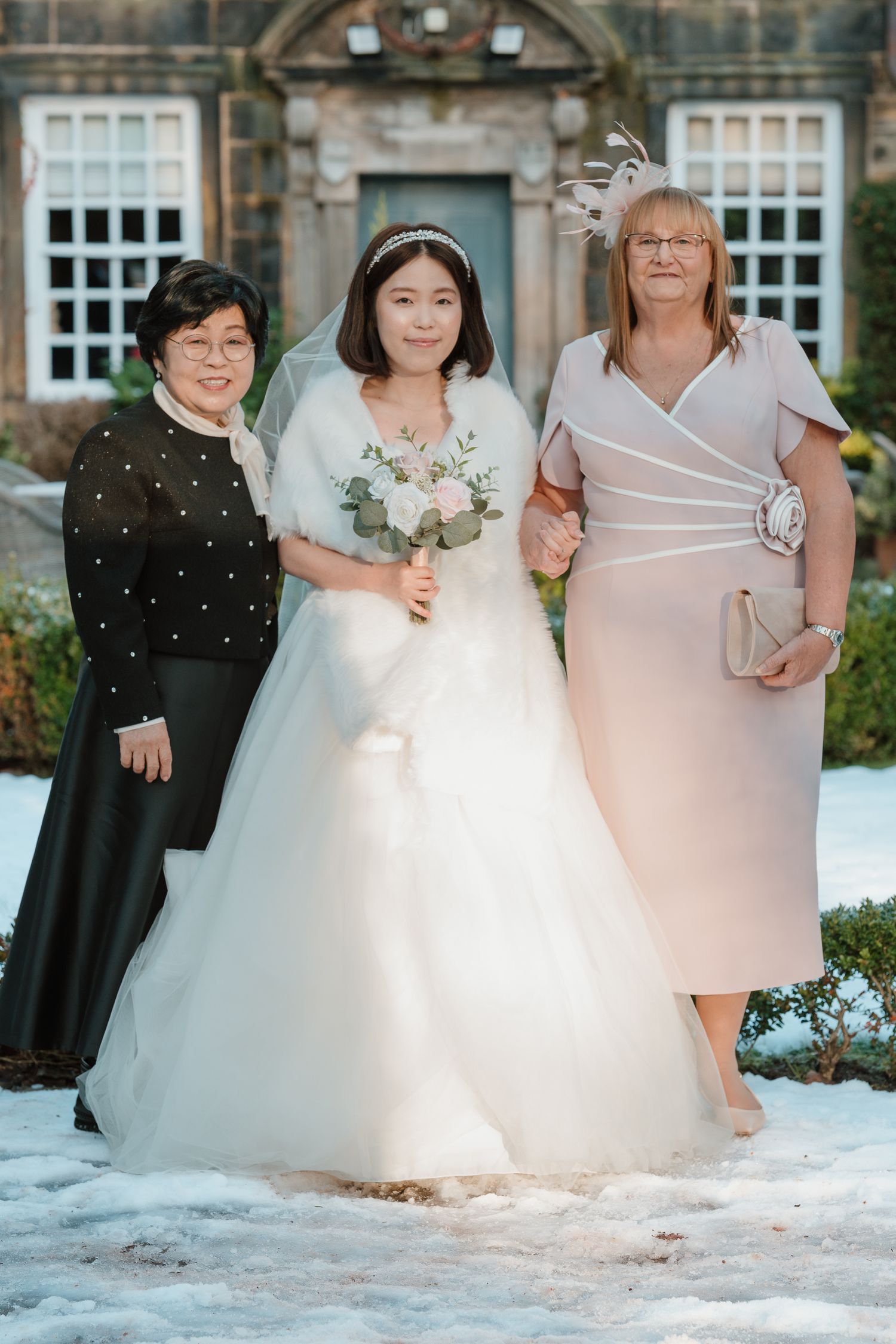 Bride in white gown and veil holds bouquet, posing with two women in formal attire outside a snowy venue. Whitley Hall Wedding