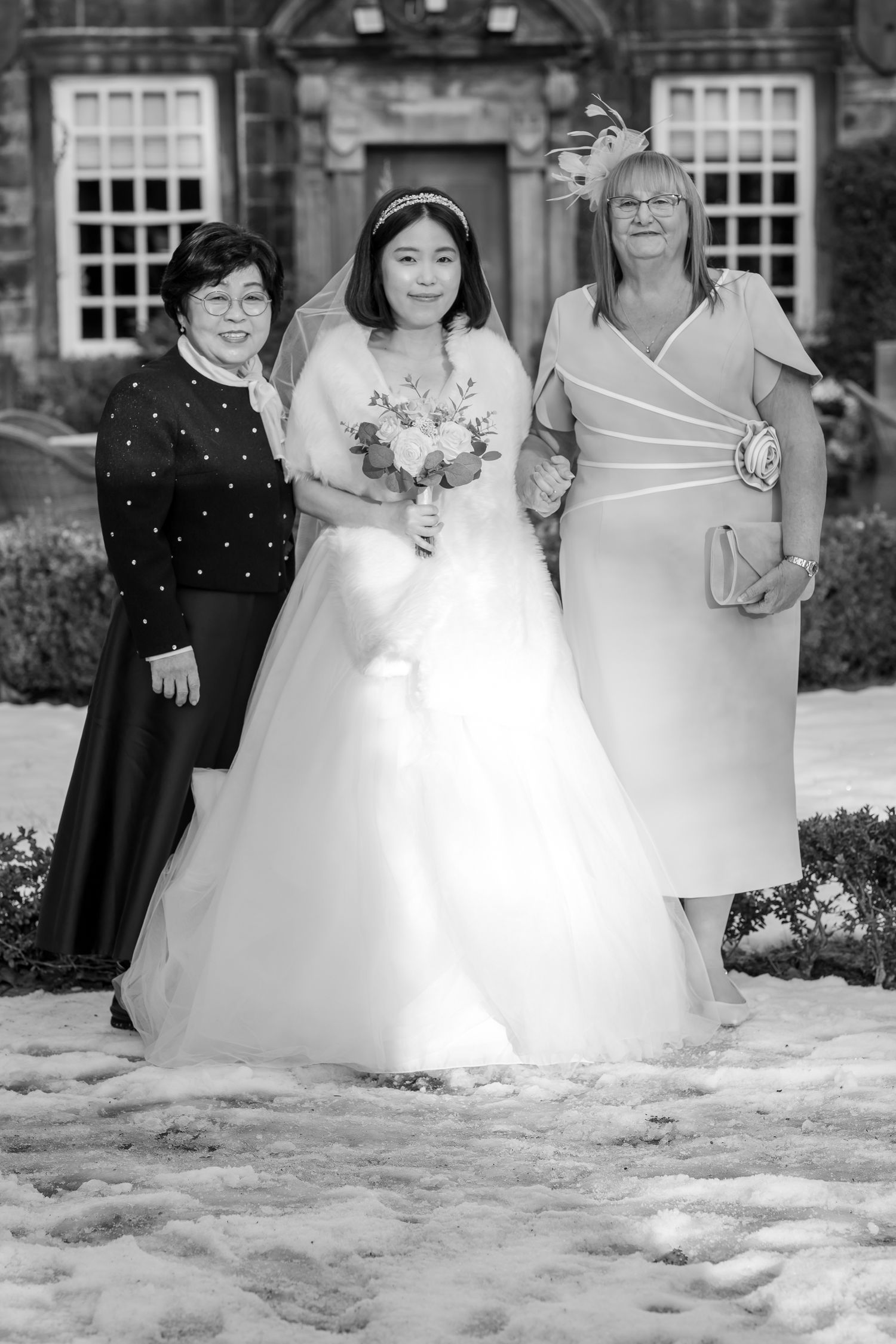 Black and white wedding photo of bride in ball gown holding bouquet, flanked by two women outside a manor.