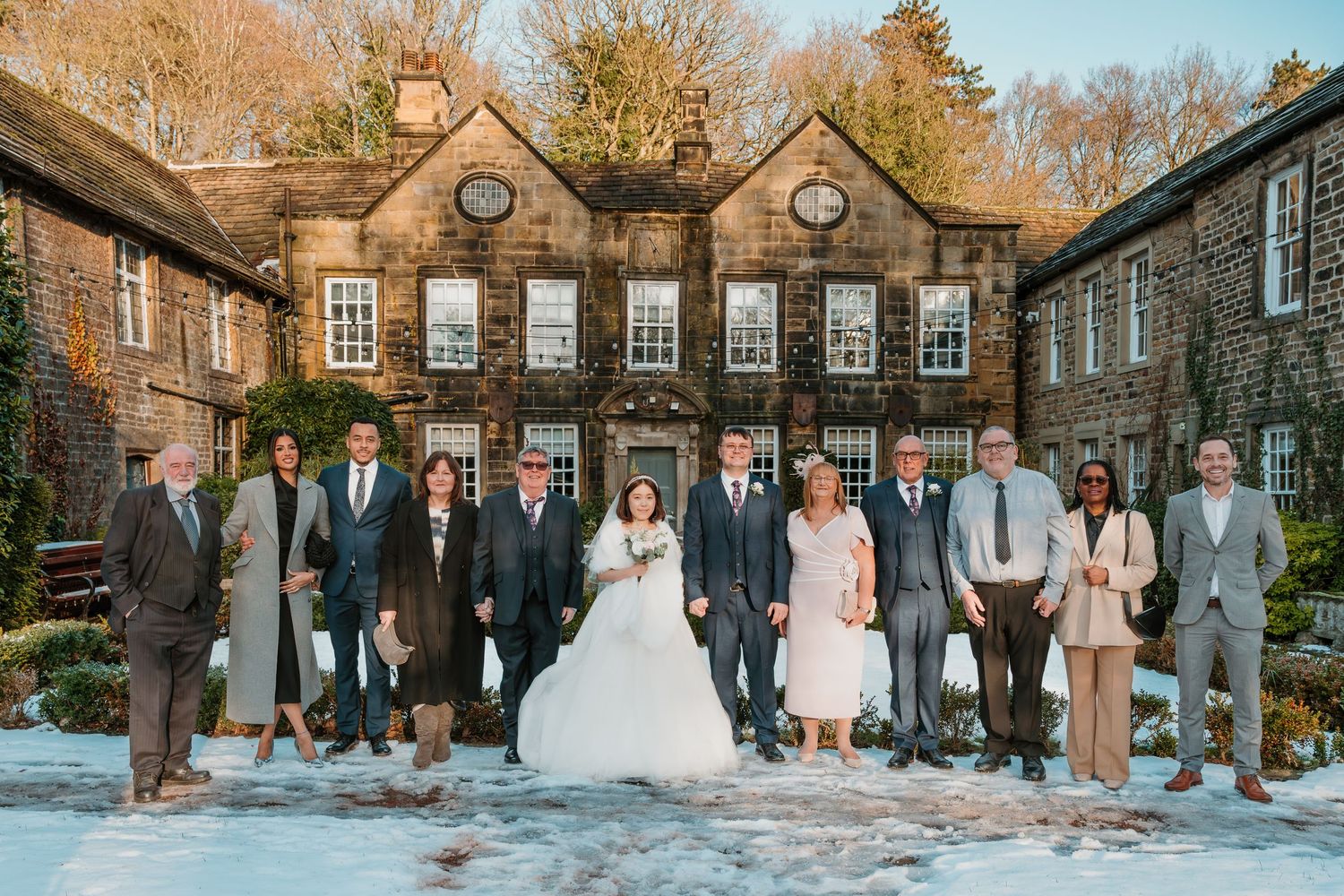 Wedding party posing in snow outside a historic stone manor house in winter, bride in white gown surrounded by guests in formal attire.
