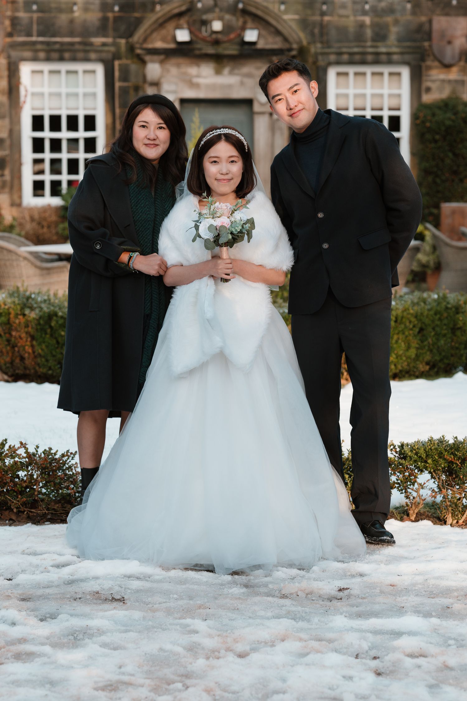Bride in white gown with fur wrap holds bouquet, flanked by two guests in black, posing in snowy garden. Whitley Hall Wedding