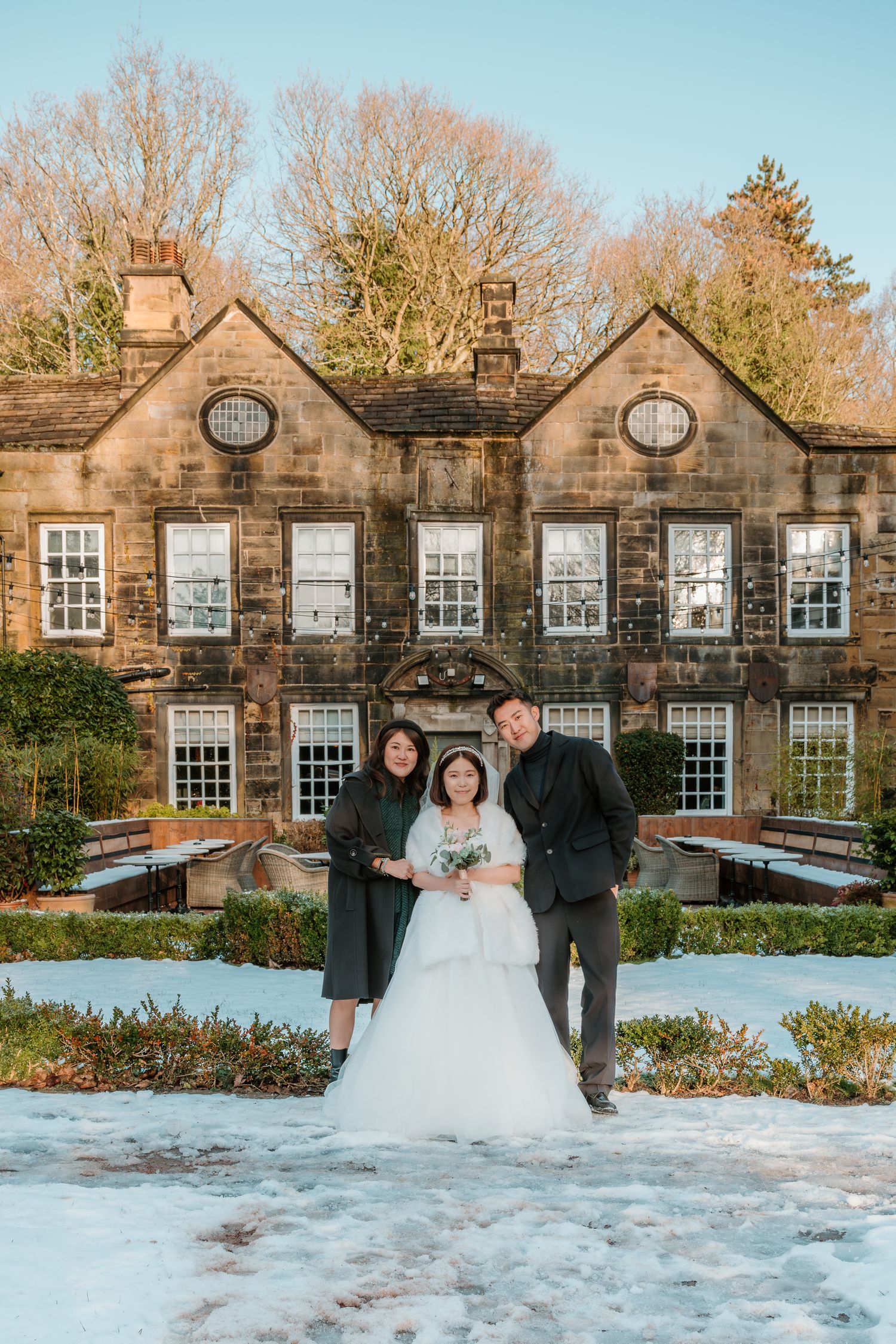 Bride and groom pose with guest outside a snow-covered stone manor house on a sunny winter wedding day.