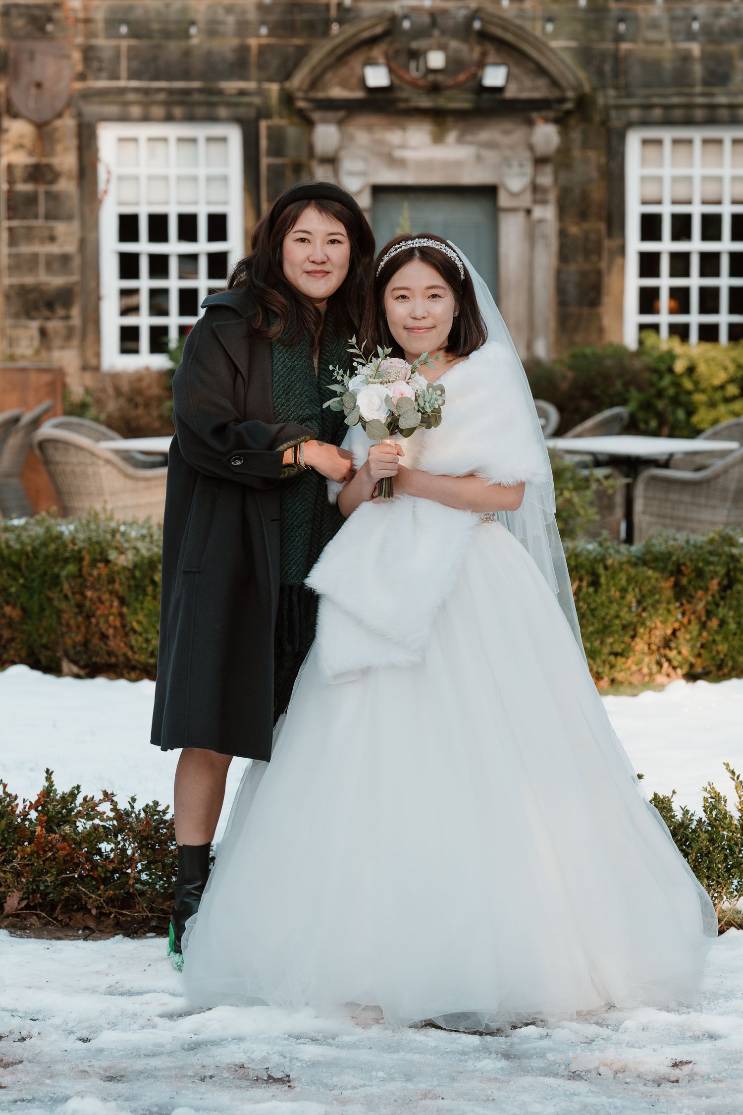 Bride in white gown holding bouquet poses with guest in dark coat on snowy winter wedding venue grounds.