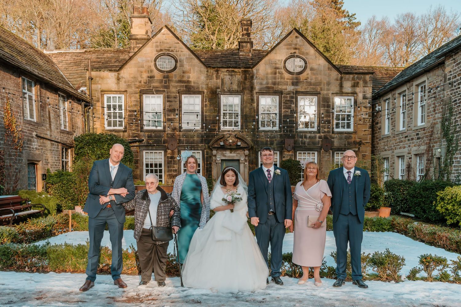 Wedding party posing in snow outside a historic stone manor house in winter sunshine.