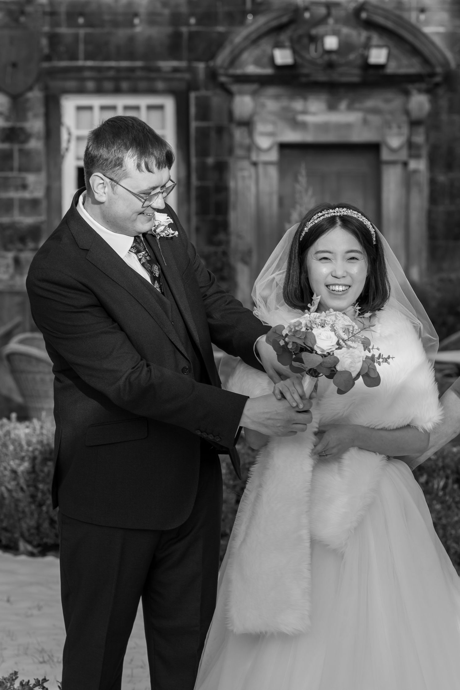 Black and white wedding photo of smiling bride in veil holding bouquet with groom in suit outside stone building.