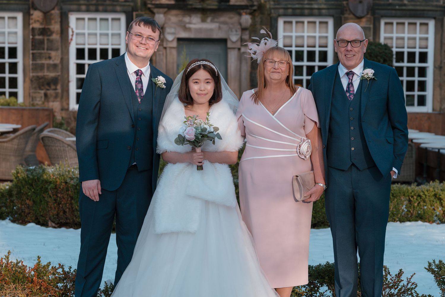 Bride in white gown with fur wrap poses with groom and parents outside snowy stone building.