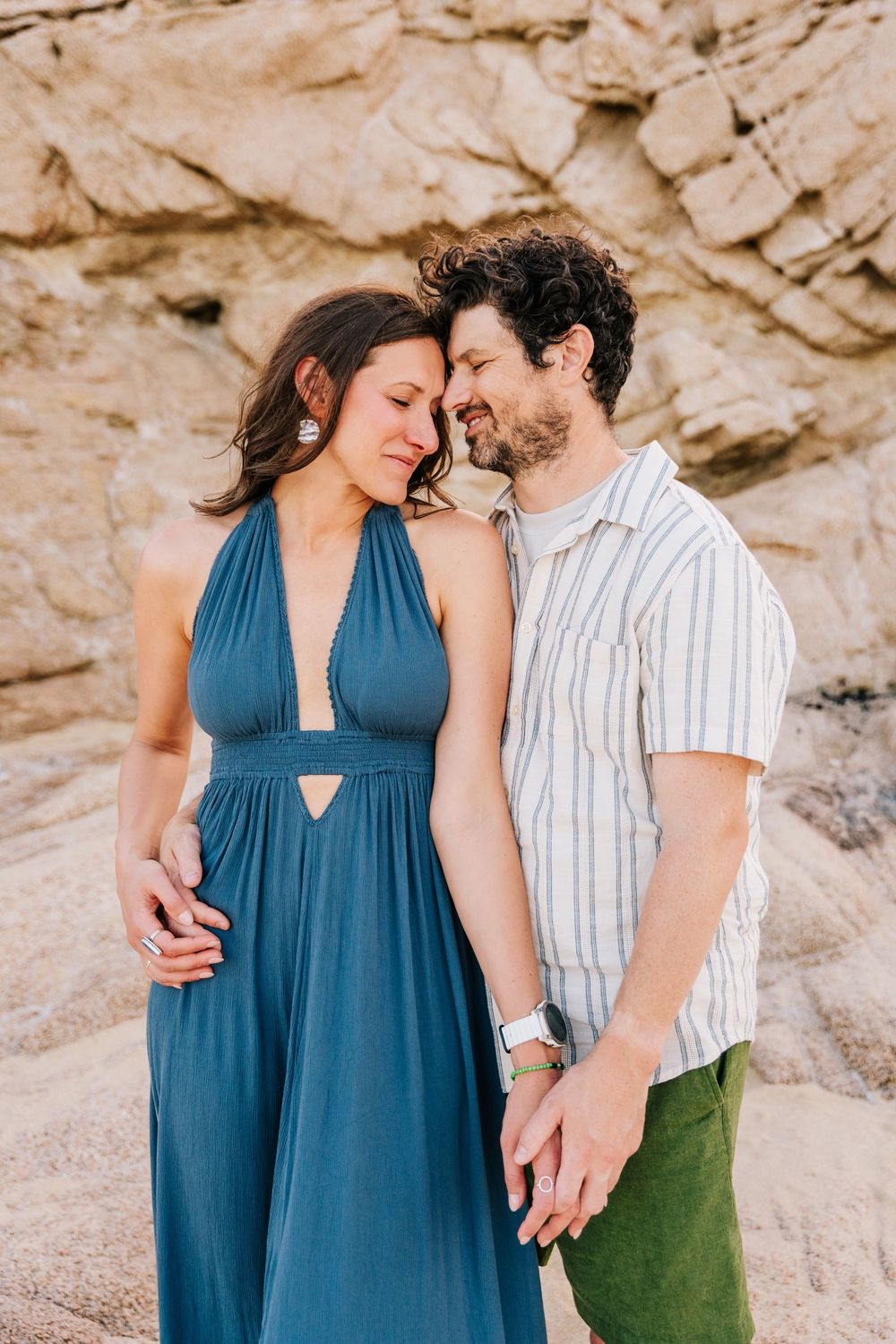 A couple shares a tender moment on a rocky beach, the woman in a teal halter dress and man in a striped shirt.