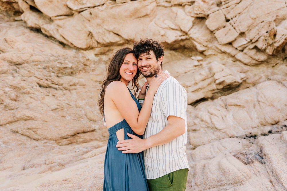 A smiling couple embraces in front of sandy rock formations, woman in blue dress, man in striped shirt and green shorts.