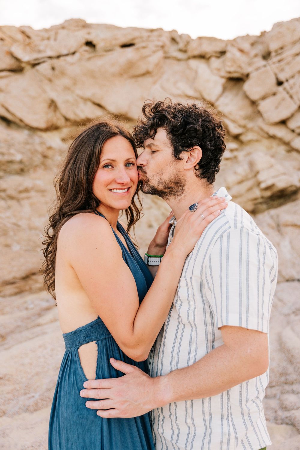 A couple embraces near sandy rocks, the man kissing the woman's temple as she smiles warmly in a teal dress.
