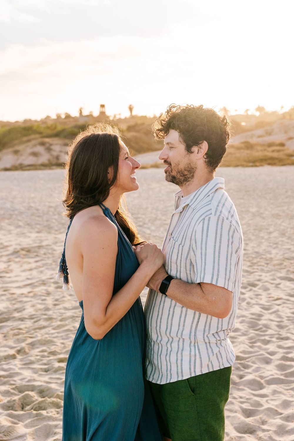 A couple shares a romantic moment on a sandy beach at golden hour, smiling at each other in casual summer attire.